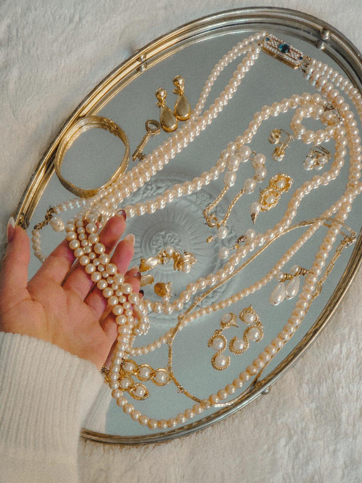 Una mano posa fili di perle, orecchini e catenine dorate su un vassoio d'argento