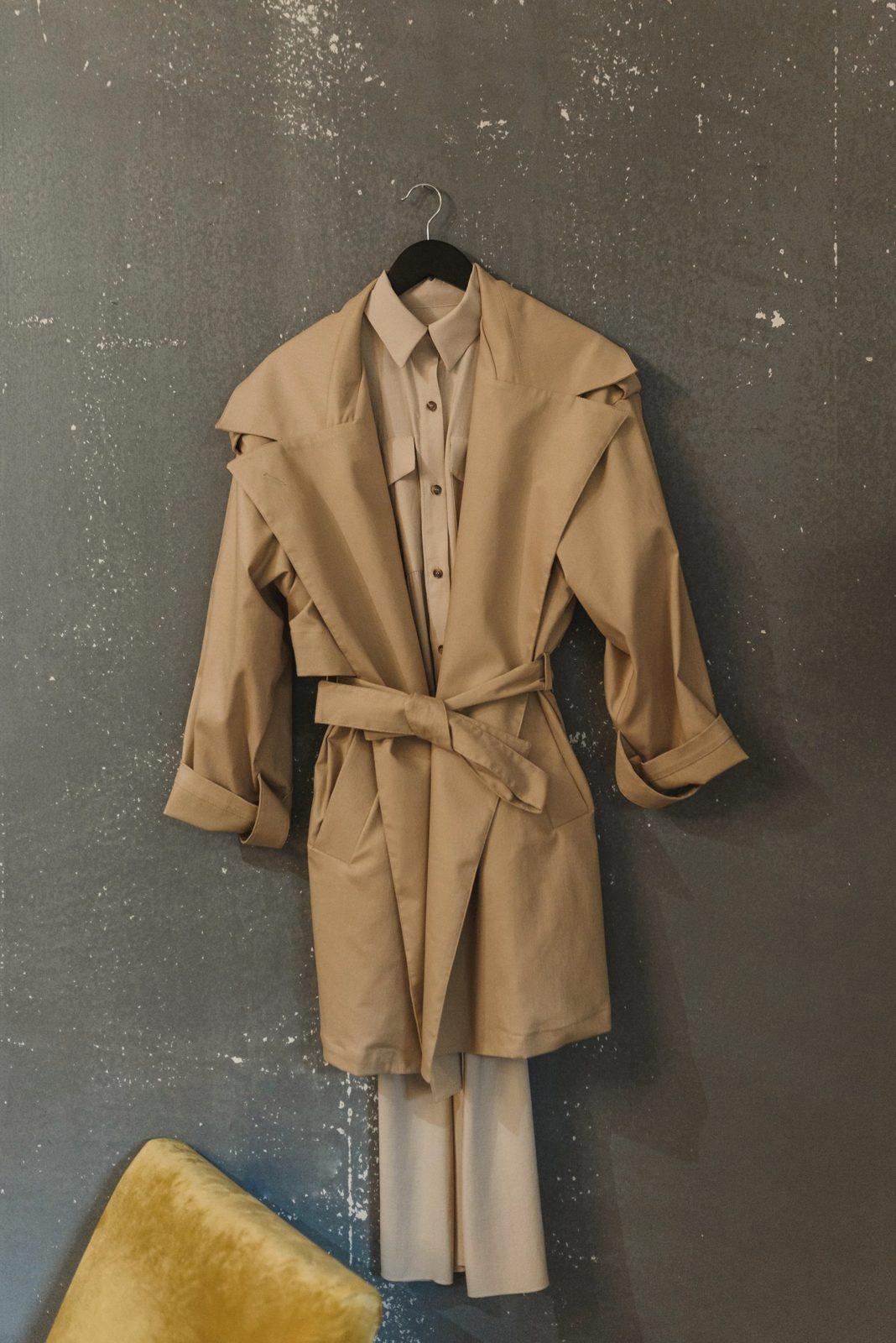 Un trench beige con la cintura annodata, appeso a una gruccia nera contro un muro grigio